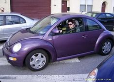 a man driving a purple car down a street next to parked cars and garage doors