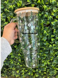 a hand holding a tumbler cup in front of a green bush with white flowers