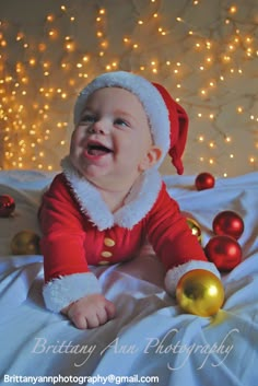 a baby wearing a santa claus outfit laying on a bed next to christmas balls and lights