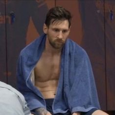 a man with no shirt sitting on a bed wrapped in a blue towel and looking at the camera