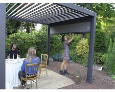 two women are sitting at a table under a pergolated roof and one woman is reaching up to the sky