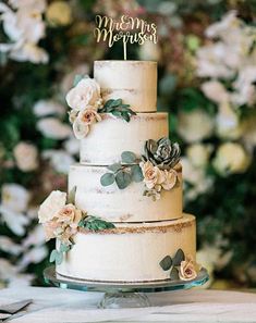 a three tiered wedding cake with flowers on the side and greenery behind it