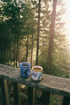 two cups of coffee sitting on top of a wooden table in front of some trees