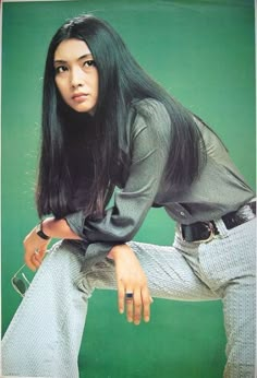 a woman with long black hair sitting on top of a green background wearing white pants