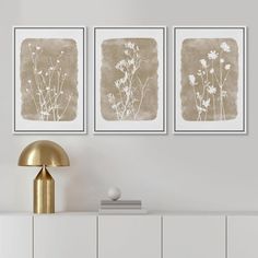 three framed art prints on a wall above a white dresser with a gold lamp and side table