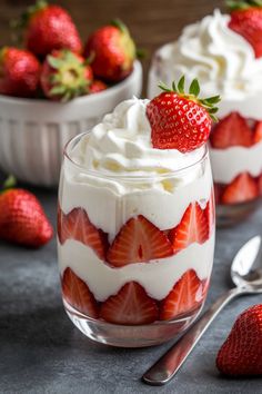 two dessert dishes with strawberries and whipped cream in them on a gray surface next to spoons