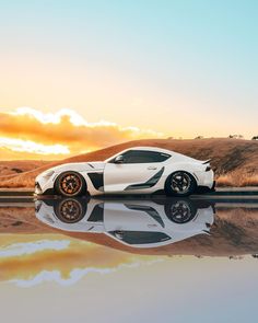 a white sports car is parked on the side of the road with its reflection in the water