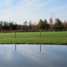 a large field with water and trees in the background, along side a fenced in area