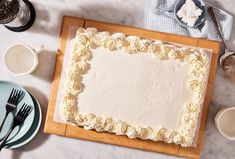 a white frosted cake sitting on top of a wooden cutting board next to cups and saucers