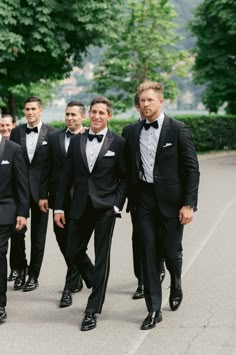 a group of men in tuxedos are walking down the street with trees behind them