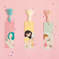 three bookmarks with tassels hanging from them on a pink surface surrounded by confetti