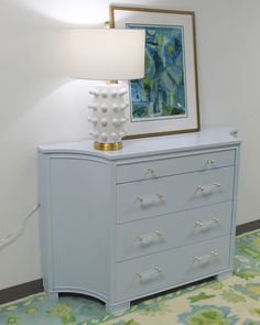 a white lamp sitting on top of a blue dresser next to a framed art piece