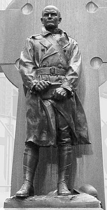 a black and white photo of a statue of a man wearing a trench coat, standing in front of a cross