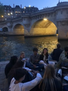 group of people sitting on the edge of a body of water near a bridge at night