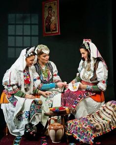 three women dressed in traditional mexican garb sitting at a small table with an urn