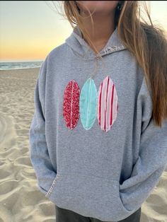 a girl standing on the beach wearing a sweatshirt with three surfboards embroidered on it