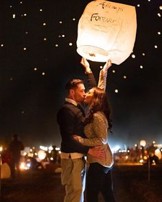 a man and woman kissing while holding up a sky lantern in the air at night