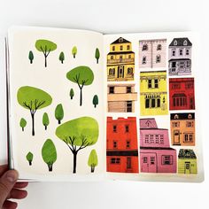 an open book with houses and trees painted on the pages, in which there is a hand holding a pencil