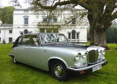 an old silver car parked in front of a large white house with trees and grass