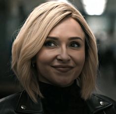 a woman with blonde hair wearing a black leather jacket and smiling at the camera while standing in front of a building