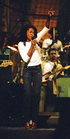 a woman standing in front of a microphone on stage with other people behind her and singing