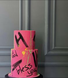 a pink and black cake with writing on the bottom layer is sitting in front of a gray wall