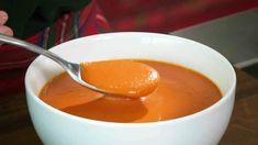 a spoon in a white bowl filled with tomato soup on a wooden table next to a person