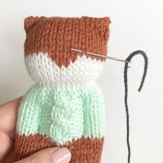 a hand holding a knitted cat ornament next to a yarn ball with a needle