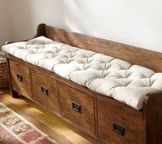 an old wooden bench with white cushions and drawers on it's sides, in front of a wall