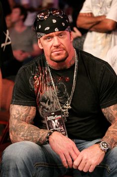 a man with tattoos sitting in front of other people at a convention or show, wearing a black t - shirt and jeans