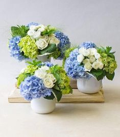 three white vases filled with blue and green flowers on a wooden stand next to each other