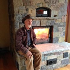 an older man sitting in front of a stone fireplace with a wood burning stove on top