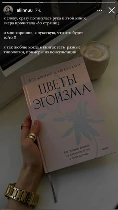 a person holding up a book on top of a table next to a cup of coffee