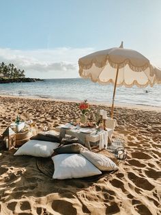 an umbrella and some pillows on the sand at the beach with food set out for dinner