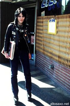 a woman standing in front of a building with her hands in her pockets and holding a book