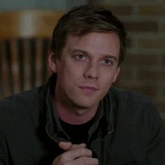 a man sitting down with his hand on his chest and looking at the camera while wearing a black shirt