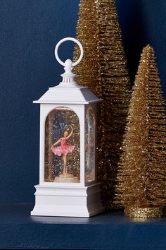 a small white lantern with a ballerina figure in it next to two christmas trees