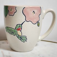 a coffee cup with a hummingbird painted on it's side and pink flowers in the background