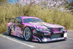 a purple car parked in a parking lot next to some bushes and trees with pink flowers on it