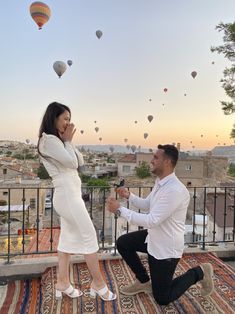 a man kneeling down next to a woman on top of a rug with hot air balloons in the sky