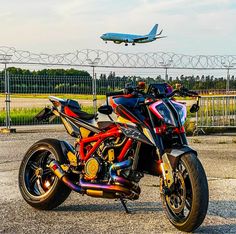 a red and black motorcycle parked in front of a fence with an airplane flying overhead