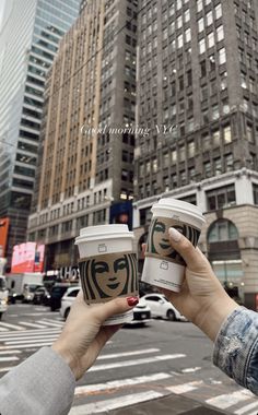 two people holding up coffee cups in the middle of a street with tall buildings behind them