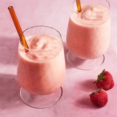 two glasses filled with smoothie and strawberries next to each other on a pink surface