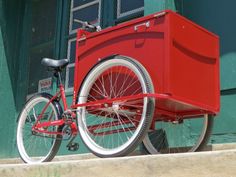 a red bike with a cooler attached to it's front wheel parked in front of a green building