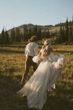 a bride and groom are walking through the grass in an open field with mountains in the background