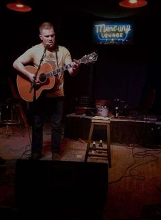 a man standing on top of a stage with a guitar in his hand and a sign above him