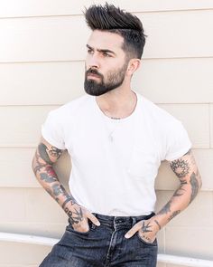 a man with tattoos and piercings standing in front of a house wearing jeans, a white t - shirt and black shoes