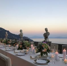 an outdoor dining table with candles and flowers on it, overlooking the ocean at dusk