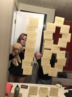 a woman holding a stuffed animal in front of a mirror with sticky notes on it