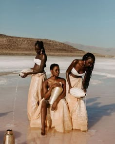 three women in white dresses are sitting on the beach and drinking water from a faucet